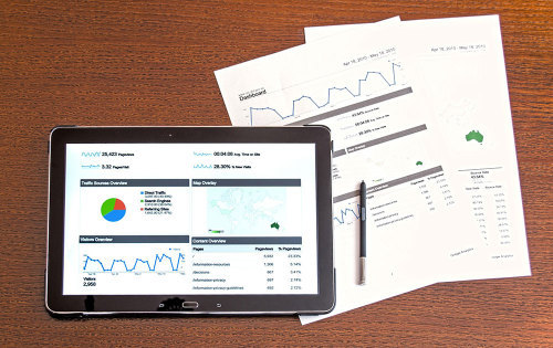 A Tablet Device Showcasing Charts And Graphs With Paperwork On The Desk To Match It
