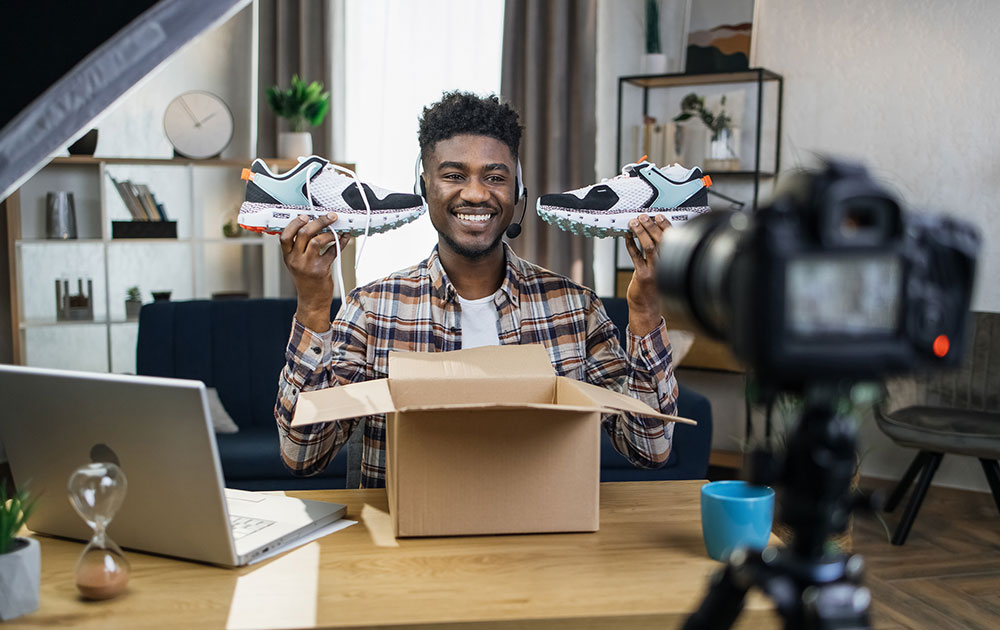 Smiling black man sitting at table with new sneakers in hands and recording video review on professional camera.